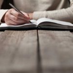 A person writing in a notebook on top of a wooden table.