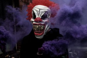 A clown mask is in front of purple smoke.